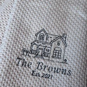 Peut inclure: Un torchon de cuisine beige à tissage gaufré avec une broderie noire représentant une maison. Sous la maison, le texte "The Browns" est suivi de "Est. 2021" en police noire. Le tissu présente un motif texturé en forme de grille.