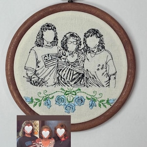 May include: A circular wooden embroidery hoop containing a black thread outline of three people standing side-by-side. Below the figures, there is an embroidered floral design featuring blue roses and green leaves on a cream-colored fabric background.