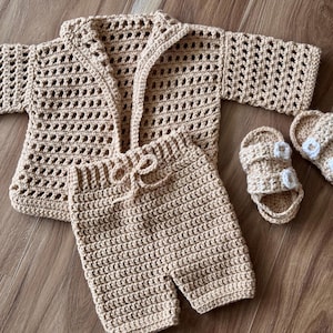 May include: A beige crochet baby outfit set, including a jacket, shorts, and sandals. The jacket has an open front and long sleeves. The shorts have a drawstring waist. The sandals have white flower accents.