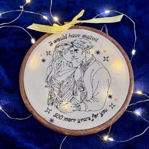 Peut inclure: Broderie sur tambour représentant un couple s'embrassant, avec le texte "I would have waited 500 more years for you." Le motif est brodé en fil noir sur fond crème, dans un cercle en bois. Un ruban jaune et des guirlandes lumineuses décorent l'ensemble.