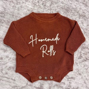 May include: A rust-colored knitted baby romper with long sleeves and a round neckline. The words "Homemade Rolls" are embroidered in white script across the front. Three wooden buttons fasten the bottom.