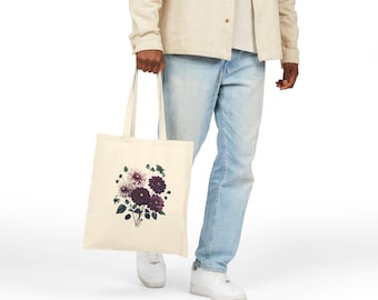 May include: Cream-colored tote bag featuring a floral design with dark purple and white dahlias and green leaves. The bag is held by a person wearing light-wash jeans and a cream-colored jacket.