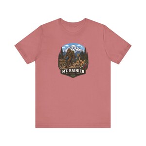 May include: A dusty rose short-sleeve t-shirt featuring a graphic of a hiker, a dog, and a Bigfoot figure walking on a trail. The background shows a snow-capped mountain and pine trees. The text reads Mt. Rainier National Park.