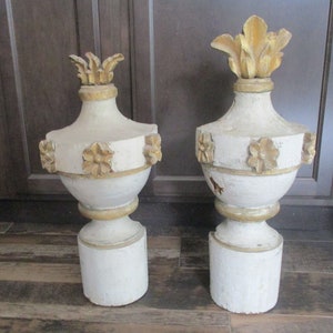May include: A pair of antique, white and gold painted wooden finials with ornate floral details. The finials are in a classic urn shape with a round base.