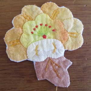 May include: A hand-stitched fabric flower with yellow, orange, and white petals. The center of the flower is white with a green and red embroidered design.