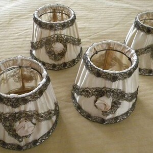 May include: Four white lampshades with lace trim and a floral design. The lampshades are made of fabric and have a delicate, vintage look.