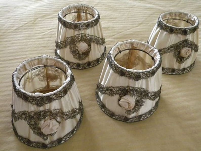 May include: Four white lampshades with lace trim and a floral design. The lampshades are made of fabric and have a delicate, vintage look.