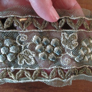 May include: A close-up of a delicate, light green lace trim with gold and silver embroidery. The trim features a repeating pattern of small flowers and leaves.