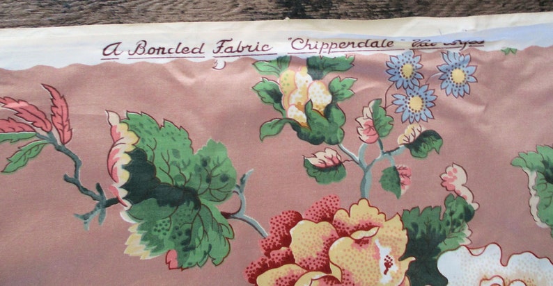 May include: A pink fabric with a floral pattern featuring green leaves, yellow and pink flowers, and blue flowers. The fabric has a label that reads "A Boncled Fabric 'Chippendale'  the  eye"