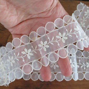 May include: White lace trim with scalloped edges and embroidered flowers. The trim features a delicate floral design and a row of small circles.