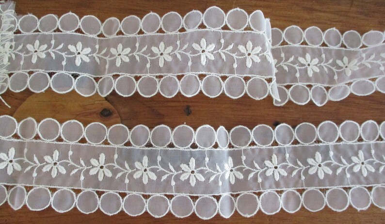 May include: Two pieces of white lace trim with scalloped edges and embroidered floral designs.