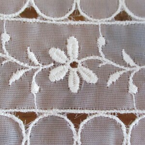 May include: White lace trim with a delicate floral pattern. The trim features small white flowers with five petals and a central hole, connected by thin, vine-like stems. The lace is woven on a sheer, translucent fabric.