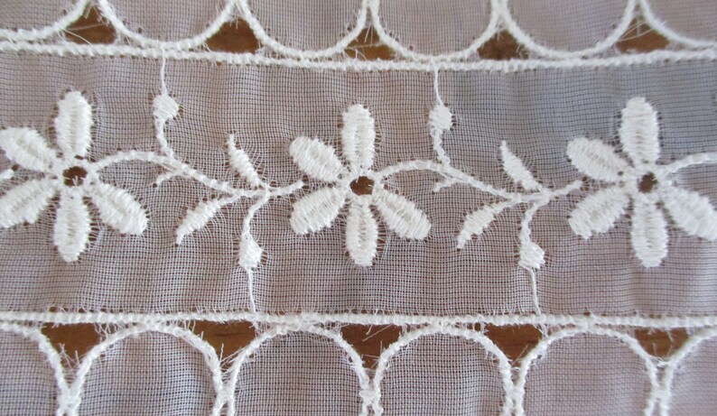 May include: White lace trim with a delicate floral pattern. The trim features small white flowers with five petals and a central hole, connected by thin, vine-like stems. The lace is woven on a sheer, translucent fabric.