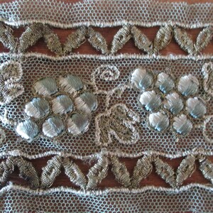 May include: A close-up of a delicate, light blue lace trim with silver and gold embroidery. The trim features a repeating pattern of stylized flowers and leaves.
