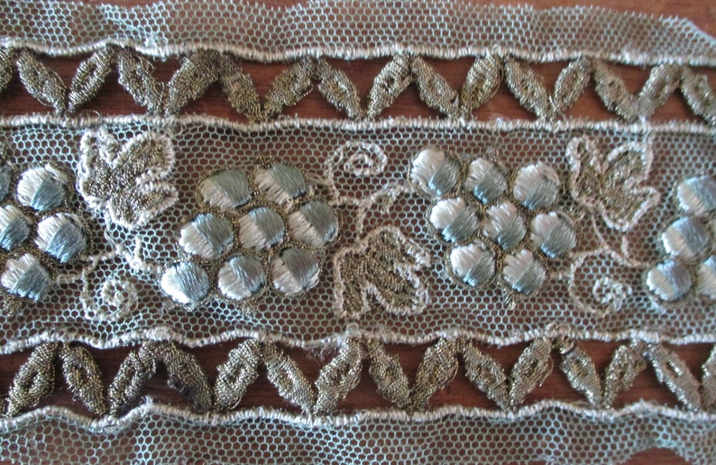 May include: A close-up of a delicate, light blue lace trim with silver and gold embroidery. The trim features a repeating pattern of stylized flowers and leaves.