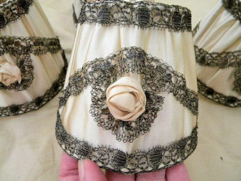 May include: Three white lampshades with black lace trim and a white fabric rose accent. The lampshades are gathered at the top and bottom.