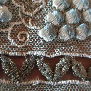 May include: Close-up of a delicate, light blue and silver lace fabric with a scalloped edge. The lace features a repeating pattern of small flowers and a beaded trim.