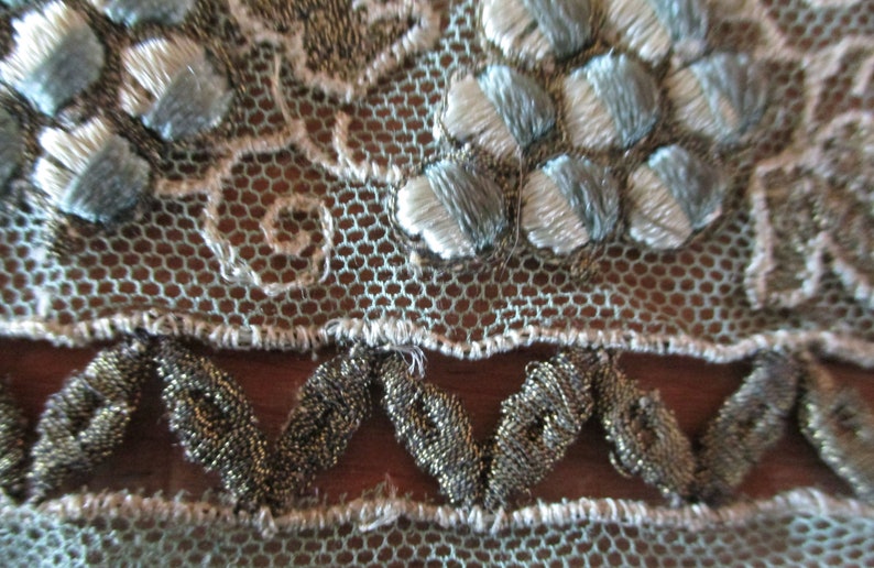 May include: Close-up of a delicate, light blue and silver lace fabric with a scalloped edge. The lace features a repeating pattern of small flowers and a beaded trim.