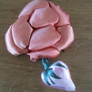 Könnte beinhalten: Eine handgefertigte, pfirsichfarbene Satinrose mit Knospe. Die Rose hat mehrere Blütenblätter und einen grünen Stiel. Die Knospe ist rosa mit grünen Blättern. Die Blume wird wahrscheinlich für Bastelarbeiten oder als Verzierung verwendet.