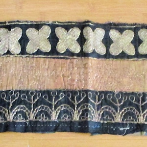 以下が含まれることがあります： 黒を基調とし、金とピンクのアクセントが施された装飾的なテキスタイルストリップ。上端と下端には金の刺繍模様があり、中央部分は明るいピンク色です。上端には金の十字模様が施されています。