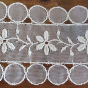 May include: White lace trim with a scalloped edge and a delicate floral design. The trim features a repeating pattern of small white flowers with a central circle.
