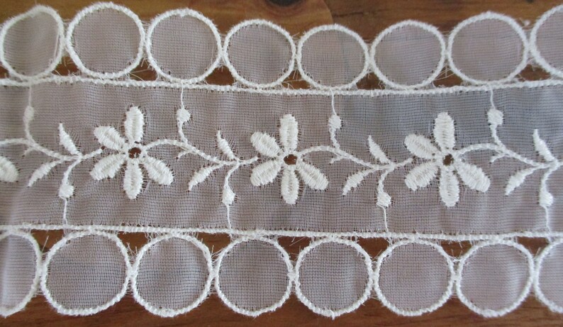 May include: White lace trim with a scalloped edge and a delicate floral design. The trim features a repeating pattern of small white flowers with a central circle.