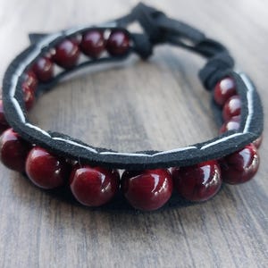 May include: A dark red wooden bead bracelet with a black leather band. The bracelet has a white stitching detail on the leather band.