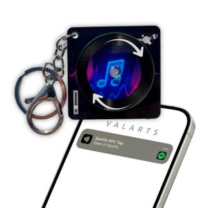 Pode incluir: Um porta-chaves preto e prateado com um design de gira-discos e um gráfico de nota musical. O porta-chaves está preso a um mosquetão e corrente prateados. Um smartphone exibe uma notificação Spotify NFC Tag.