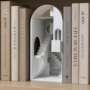 Op de afbeelding: Een witte architectonische boekensteun met een trapontwerp, geplaatst tussen verschillende boeken op een houten plank. De boeken hebben titels als "AD at 100", "The Kinfolk" en "Live Beautiful". De boekensteun voegt een decoratief element toe.