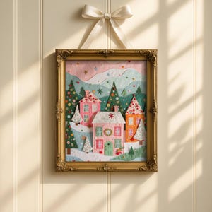 May include: A framed Christmas-themed artwork featuring a winter village scene. The artwork depicts colorful houses, snow-covered hills, and evergreen trees. The frame is ornate gold, and the artwork is suspended by a cream-colored ribbon tied in a bow.