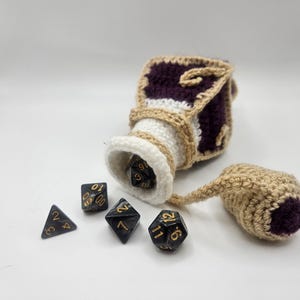A purple diamond-shaped crochet potion bottle pours polyhedral dice on to a flat surface