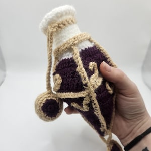 An open diamond-shaped crocheted potion bottle dice bag with gold detailing and curls and an attached golden cork with a purple top hanging to one side is held in the palm of a hand. A small charm hangs from the bottom.