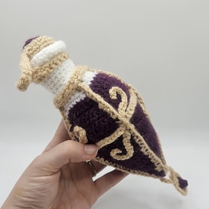 A diamond-shaped crocheted potion bottle dice bag with gold detailing and curls is held in the palm of a hand. A small charm hangs from the bottom.