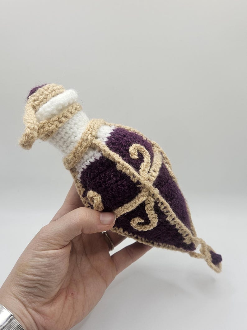 A diamond-shaped crocheted potion bottle dice bag with gold detailing and curls is held in the palm of a hand. A small charm hangs from the bottom.