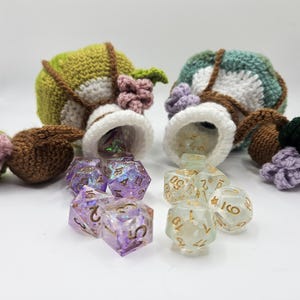 Two green crocheted potion bottle dice bag pouches spill two sets of polyhedral gaming dice onto a flat surface