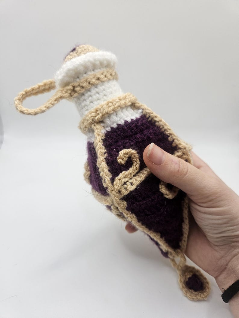 A diamond-shaped crocheted potion bottle dice bag with gold detailing and curls is held in the palm of a hand. A small charm hangs from the bottom.