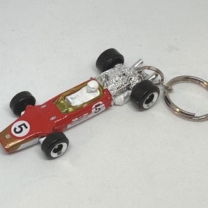 Puede incluir: Un llavero de coche de carreras vintage rojo y dorado con ruedas negras y un motor plateado. El número 5 es visible en la parte delantera del coche. El llavero incluye un llavero plateado.