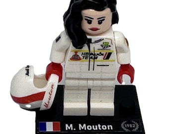 May include: A Lego minifigure of a race car driver, wearing a white racing suit with red accents and a black helmet. The figure has black hair and is standing on a black base with the name "M. Mouton" and the French flag.