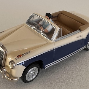 Op de afbeelding: Een vintage speelgoedcabriolet in crème en marineblauw met een bruin interieur. De auto heeft chromen accenten en een kentekenplaat. Een kleine figuur is zichtbaar achter het stuur.