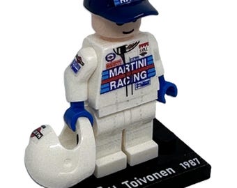 May include: A Lego minifigure of a race car driver wearing a white racing suit with blue and red Martini Racing logos, a blue cap with a Martini Racing logo, and a white helmet. The figure is standing on a black base with a Finnish flag and the text "H. Toivonen 1987".