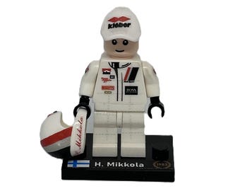 May include: A white Lego minifigure of a race car driver wearing a white racing suit with red and black accents, a white cap with the word "Kiekko" in black, and a black helmet with red and white accents. The minifigure is holding a black helmet with red and white accents. The minifigure is standing on a black base with a Finnish flag and the text "H. Mikkola 1983".