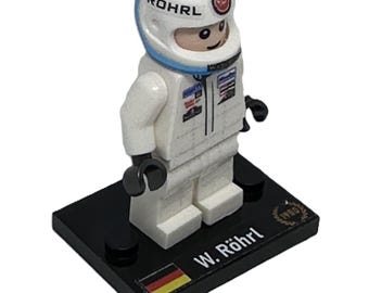 May include: A white Lego minifigure of a race car driver wearing a white helmet with the word "ROHRL" on it. The figure is standing on a black base with the text "W. Röhrl" and a German flag.