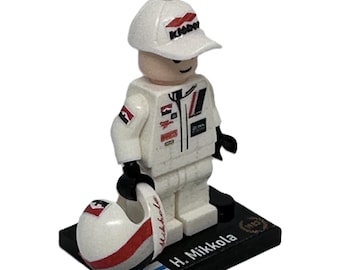 May include: A white Lego minifigure of a race car driver wearing a white racing suit with red and black accents, a white cap with a red logo, and holding a white helmet with a red stripe. The minifigure is standing on a black base with the name "H. Mikkola" and the Finnish flag printed on it.