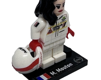 May include: A Lego figurine of a woman in a white racing suit, holding a white and red helmet. The figurine has black hair and red gloves. The base is black with the name "M. Mouton" and a French flag.