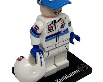 May include: A Lego minifigure of a race car driver wearing a blue and white racing suit, a blue baseball cap with the word "Michelin" on it, and a white helmet. The figure is standing on a black base with a Finnish flag and the name "J. Kankkunen".