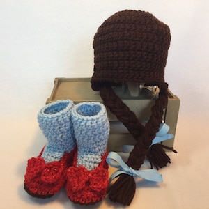May include: A set of crocheted baby booties and a hat with long braids. The booties are blue and red, and the hat is brown. The braids are brown and have blue ribbons tied at the ends.
