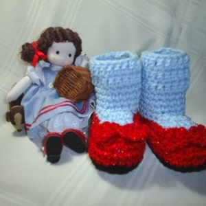 May include: A pair of blue and red crocheted baby booties with a bow on the toe. The booties are on a white surface.