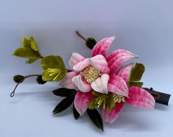 May include: A floral hair clip featuring a large pink and white flower with a detailed center, surrounded by green leaves and stems. The clip has a dark brown barrette clasp, perfect for adding a touch of nature-inspired elegance to any hairstyle.