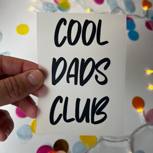 Könnte beinhalten: Eine weiße Karte mit dem Text "COOL DADS CLUB" in fetten schwarzen Buchstaben. Die Karte wird von einer Hand gehalten, mit bunten Konfetti und Lichterketten im Hintergrund.