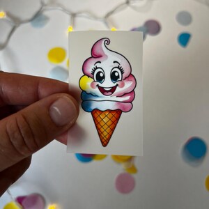 Peut inclure: Un autocollant de cornet de glace coloré avec un visage souriant. La glace est composée de couches roses, blanches, jaunes et bleues. Le cornet est orange et marron. L'autocollant est tenu sur un fond blanc avec des confettis colorés.